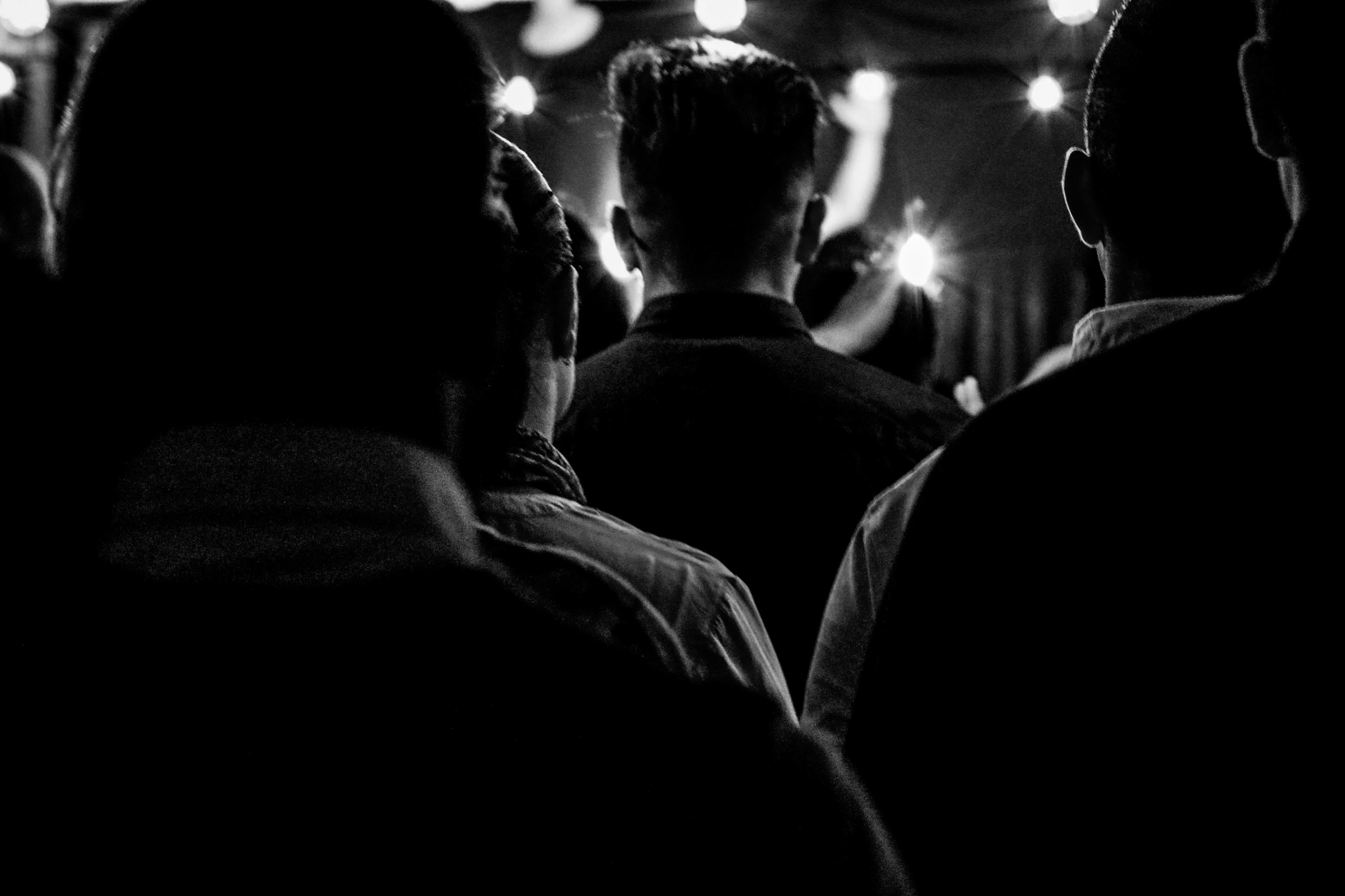 Black and white photograph taken from within an audience, showing the backs of several heads silhouetted against stage lights.